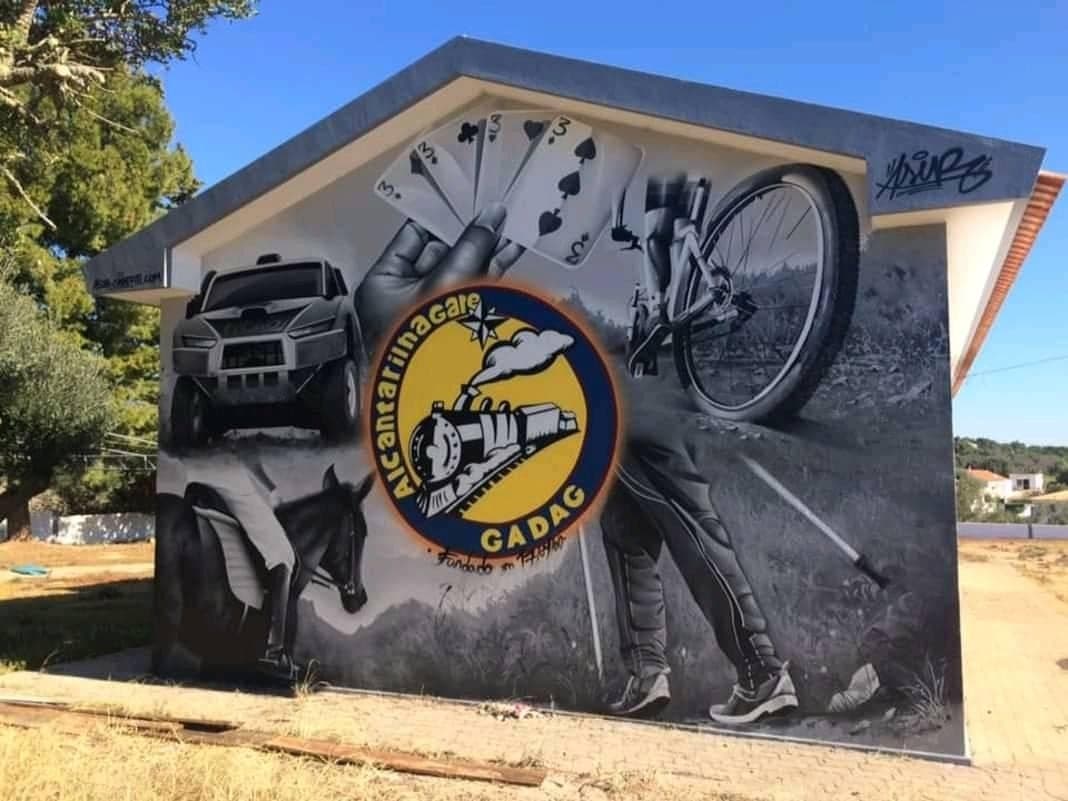 Mural pintado na fachada da sede do GADAG com o emblema do clube, um jipe, uma bicicleta, um cavalo e cartas de sueca